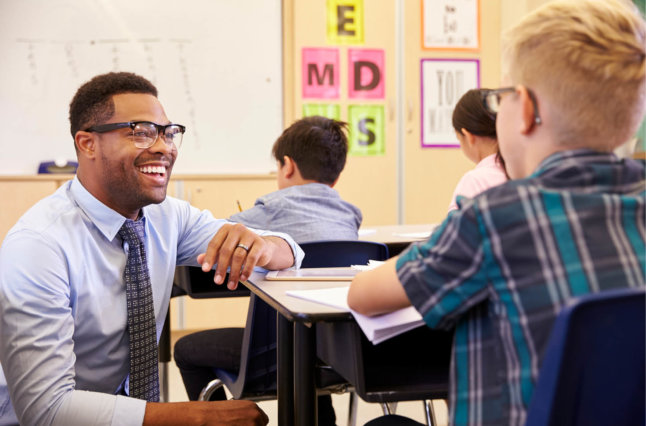 Smiling Teacher Kneeling Beside Elementary School Pupils Desk