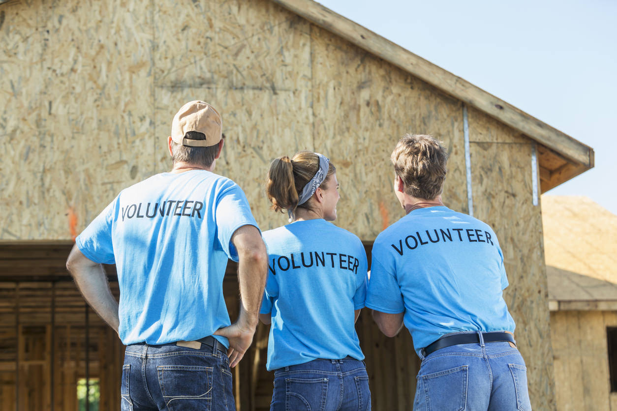 A group of three volunteers helping to build a house for a family in need. One woman stands in the middle between two men. Their backs are to the camera, showing the word VOLUNTEER written on the back of their blue shirts.