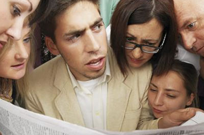 A diverse group of people huddled together reading the same newspaper