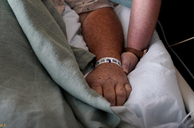 A woman holds the hand of her husband who is in the hospital suffering with COVID-19