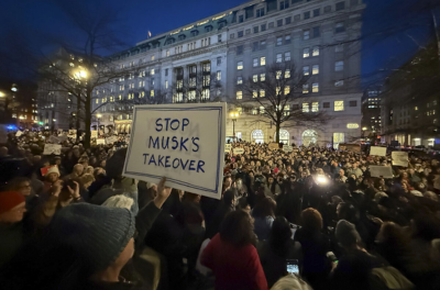 A large crowd gathers at night in front of a lit building, holding signs, including one that states "Stop Musk's Takeover," showing protest and solidarity.
